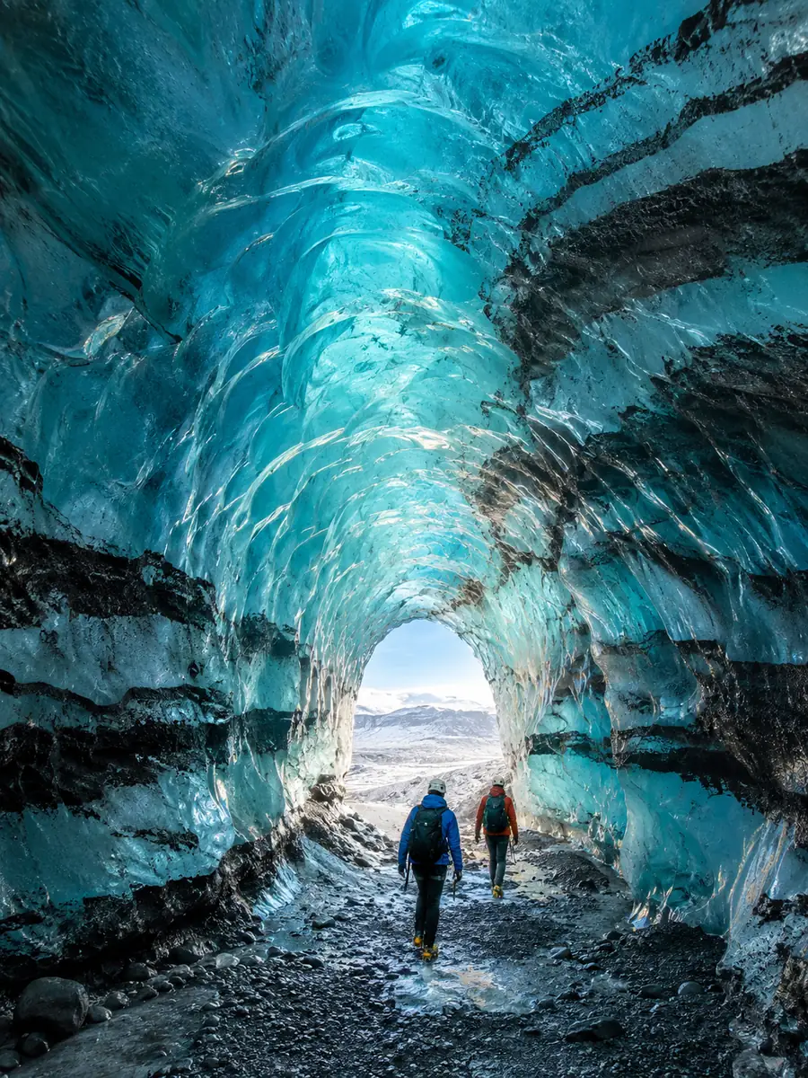 Two explorers walking out through a tunnel of blue ice with black ash layers