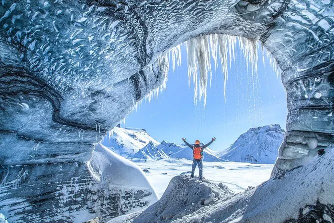 An ice cave arch opening onto snow in winter, with a person raising their arms at the entrance